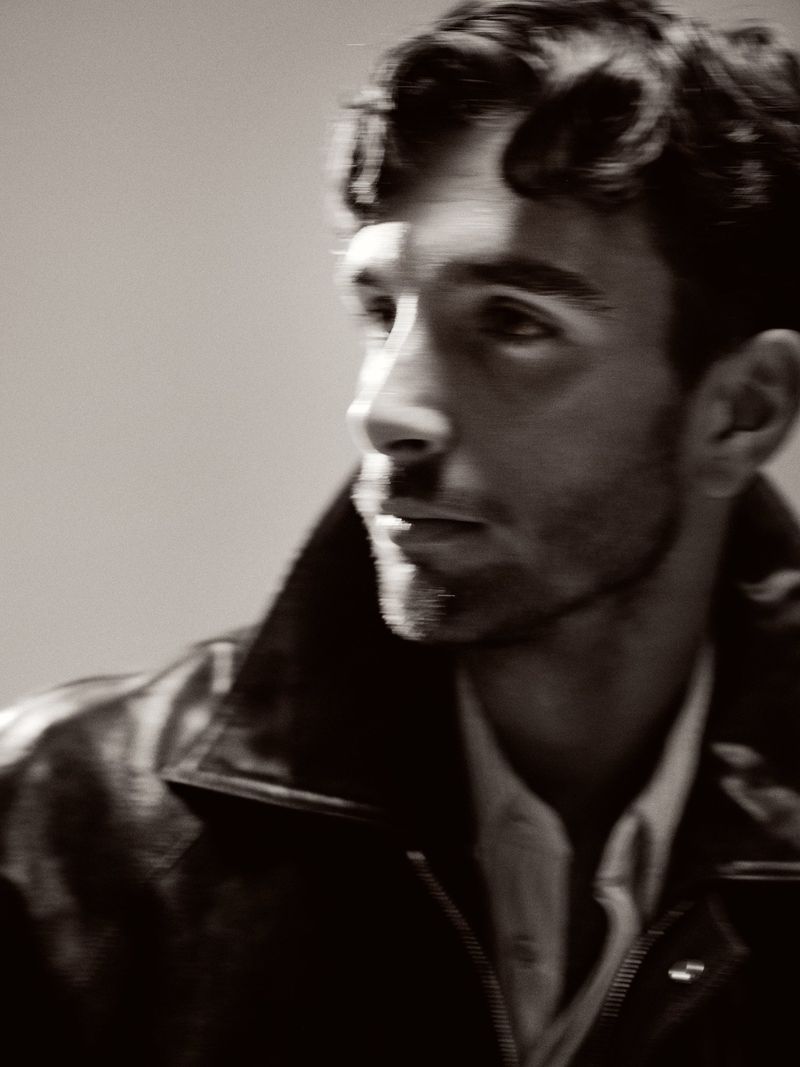 Portrait of a man with dark curly hair wearing a black leather jacket and white shirt, looking left with soft lighting.