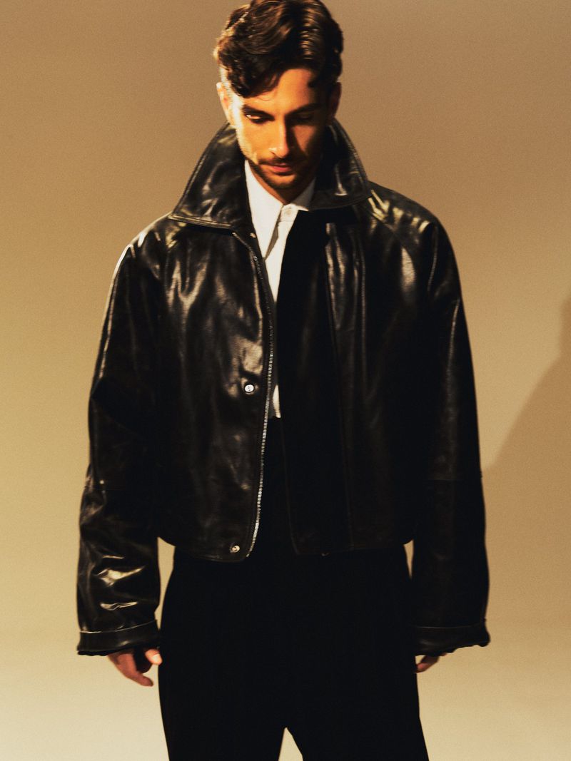 Portrait of a man wearing a leather jacket, white shirt, and black pants against a beige background in a studio setting.