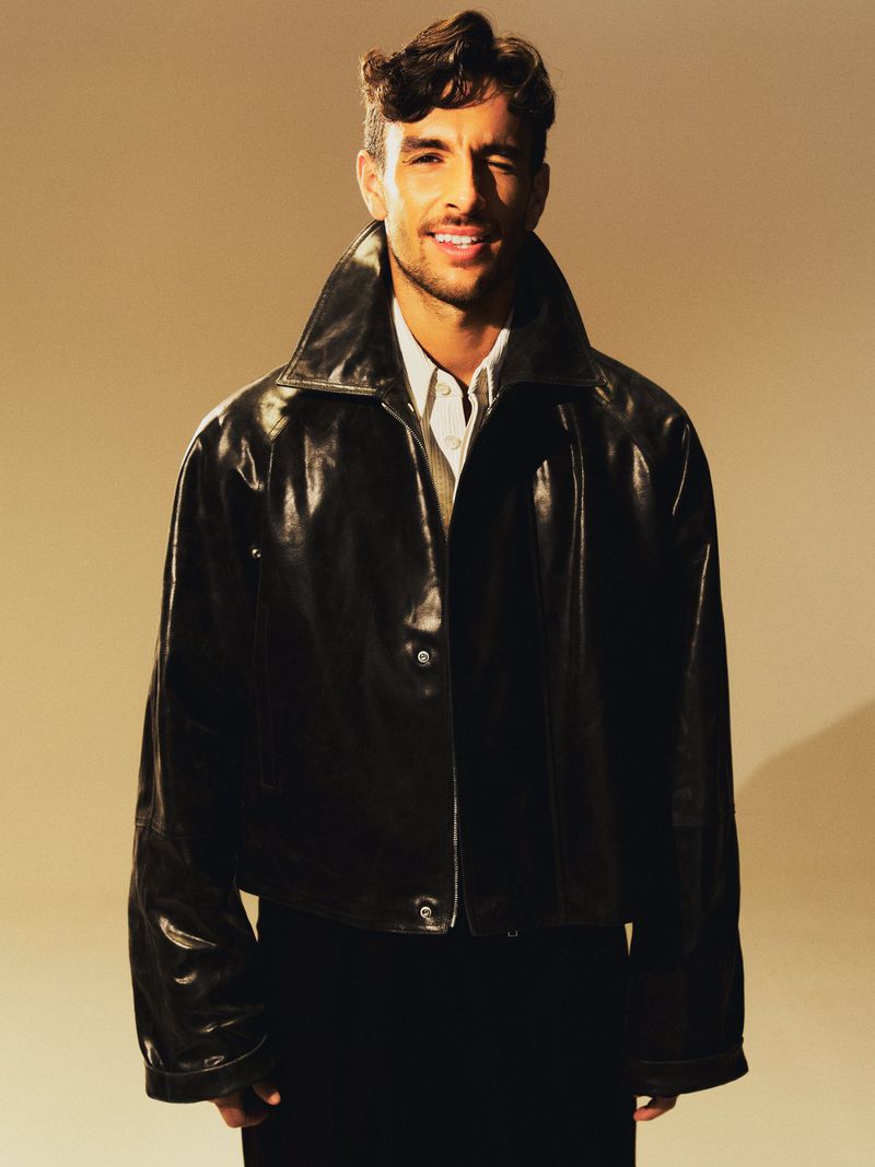Portrait of a young adult man smiling in a studio setting wearing a black leather jacket and a white collared shirt.