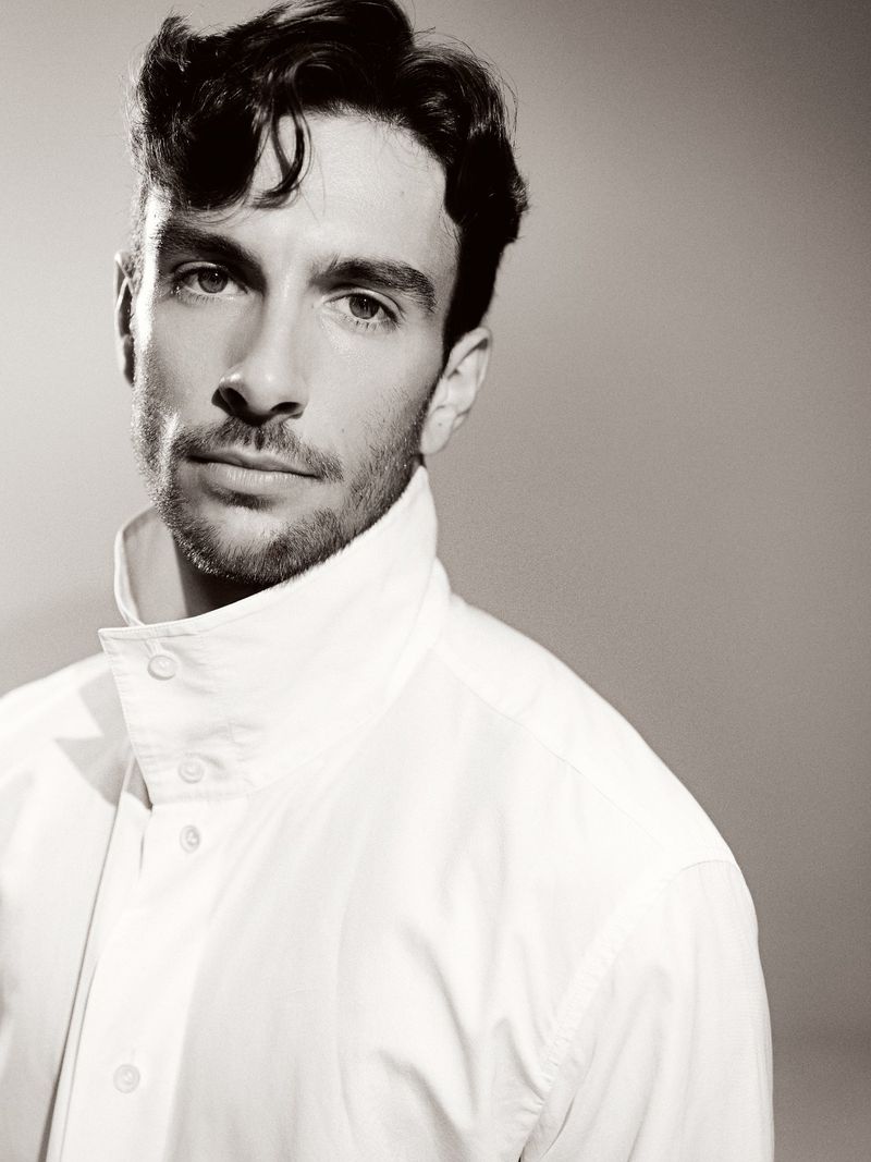 Monochromatic portrait of a bearded adult male with dark hair and a white collared shirt against a neutral grey backdrop.