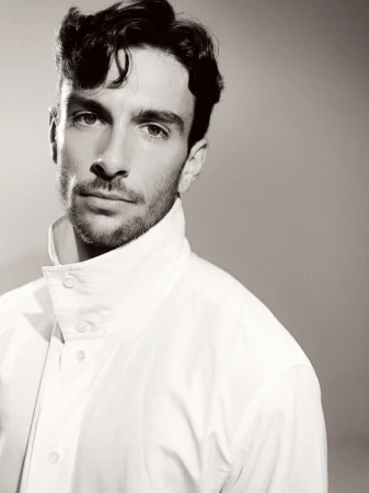 Monochromatic portrait of a bearded adult male with dark hair and a white collared shirt against a neutral grey backdrop.