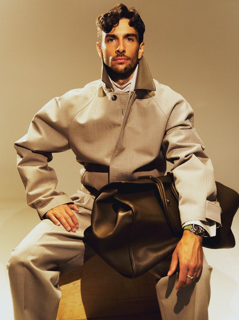 Attractive man in a grey trench coat sits holding a duffel bag. He's wearing a collared shirt, slacks, and a wristwatch in a studio setting.