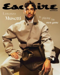 Lorenzo Musetti seated on a studio floor holding a large dark duffel bag, wearing a grey structured jacket with a high collar.