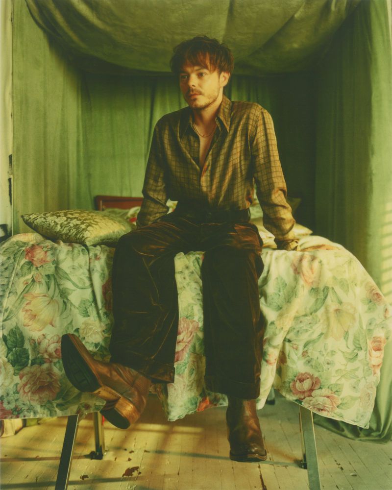 A young adult sits on a floral bed in an indoor space, clad in a patterned shirt, velvet pants, and leather boots, highlighted by natural lighting.