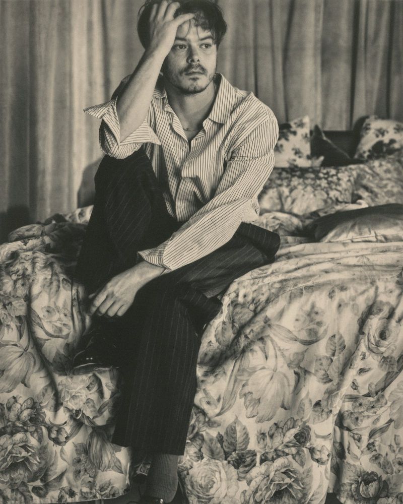 Monochrome portrait of a thoughtful man sitting on a floral bedspread, wearing a striped shirt and dark pants in a casual pose.