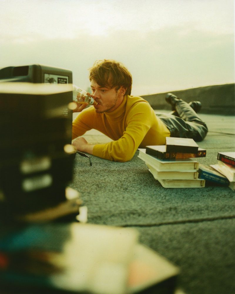 Man lying on rooftop drinking from bottle surrounded by books and a vintage television set against a bright sky in soft light.