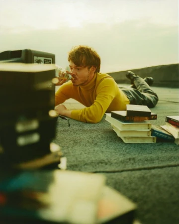 Man lying on rooftop drinking from bottle surrounded by books and a vintage television set against a bright sky in soft light.