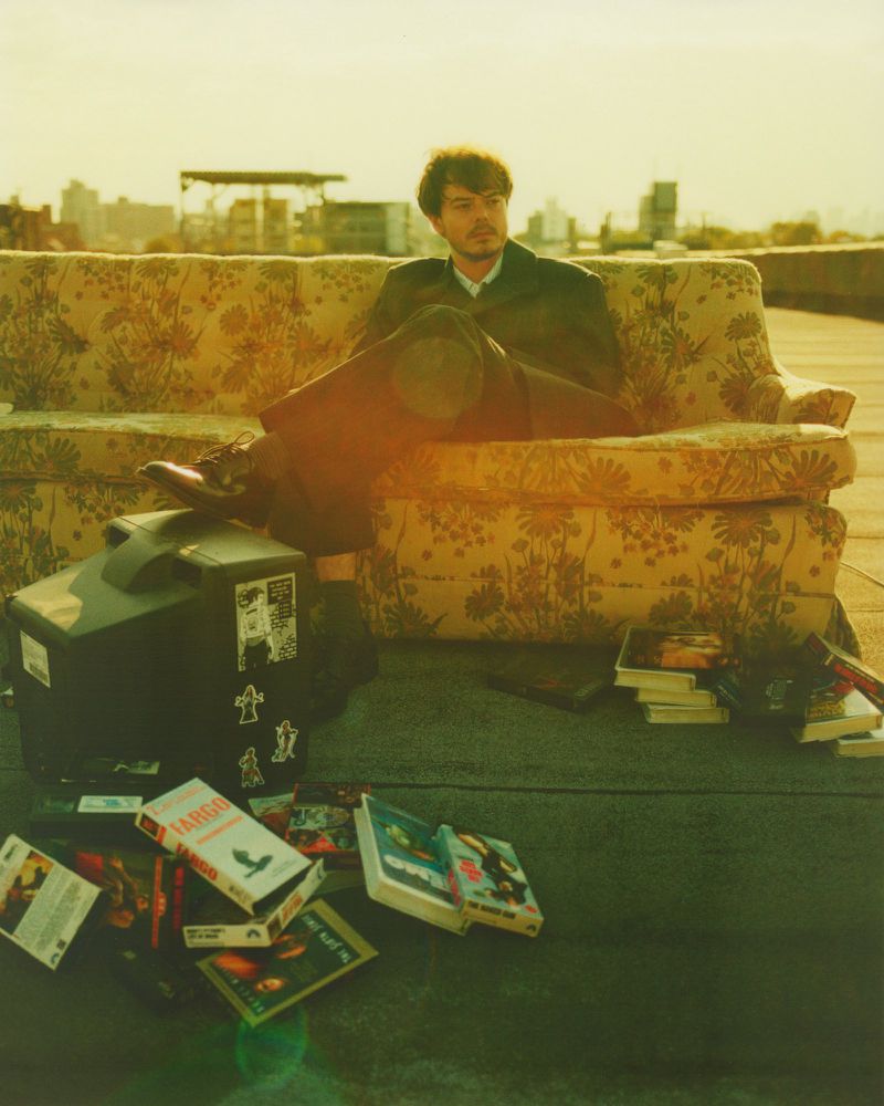 A man in a suit relaxes on a floral sofa atop a building, surrounded by VHS tapes and an old TV, with a cityscape in the background.