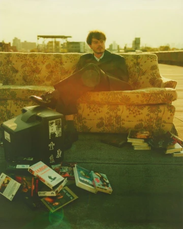 A man in a suit relaxes on a floral sofa atop a building, surrounded by VHS tapes and an old TV, with a cityscape in the background.