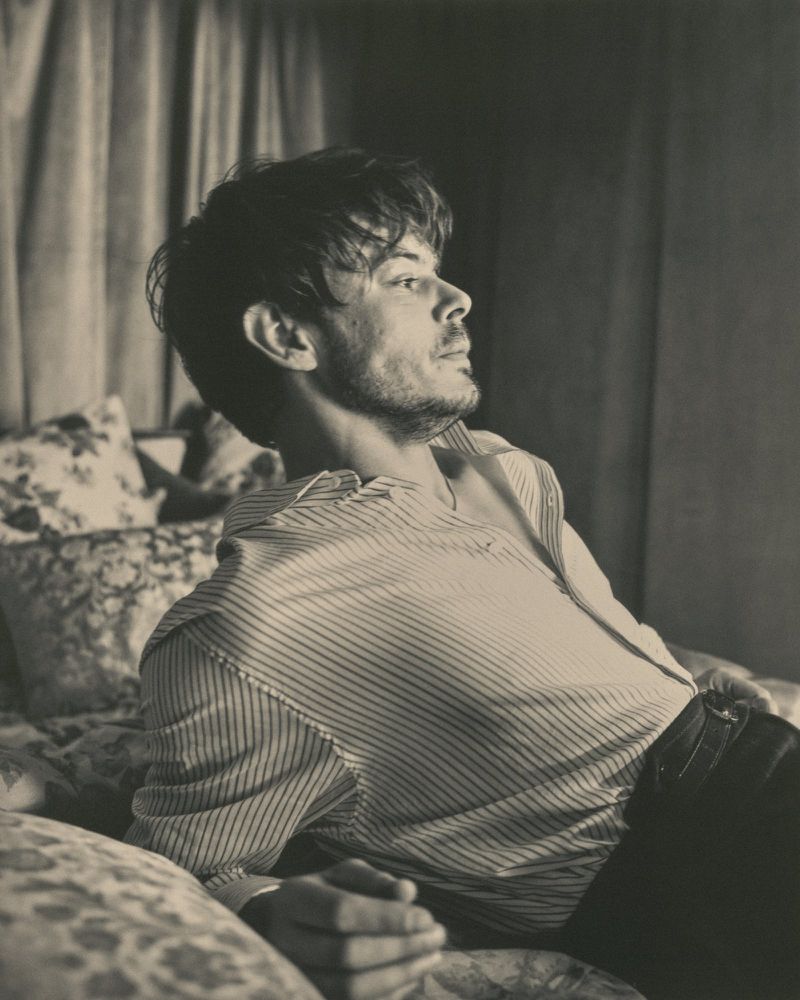 Monochrome portrait of a man with tousled brown hair reclining on a bed with floral pillows and a striped shirt, gazing toward the upper right.