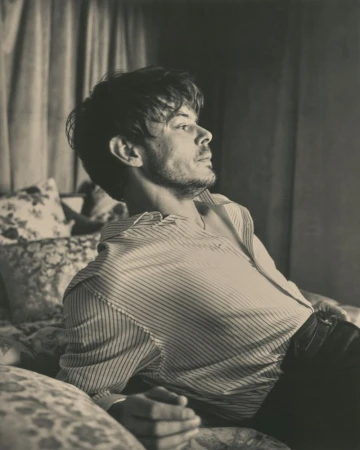 Monochrome portrait of a man with tousled brown hair reclining on a bed with floral pillows and a striped shirt, gazing toward the upper right.