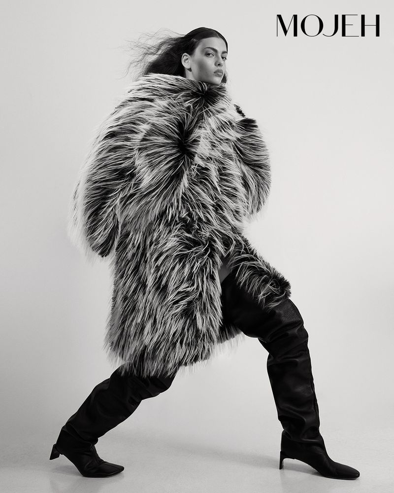 Fashion editorial shot of a woman in a fur coat and thigh-high boots striking a pose against a neutral background, showcasing modern style.