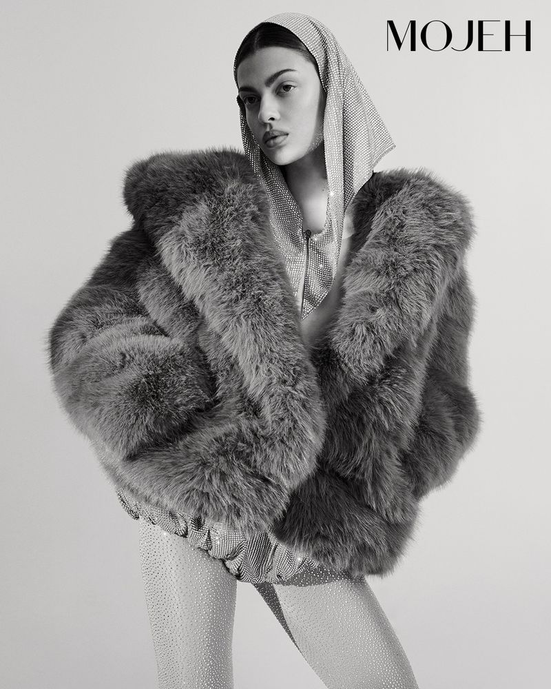 Monochrome studio shot of a woman in a fur coat, sparkling top and leggings, and a jeweled headscarf posing in front of a white backdrop.