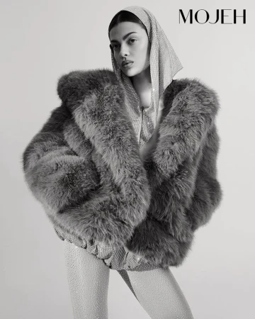 Monochrome studio shot of a woman in a fur coat, sparkling top and leggings, and a jeweled headscarf posing in front of a white backdrop.