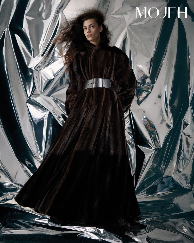 Fashion shot of a woman in a dark brown fur coat with a wide silver belt, posing in front of a shiny silver backdrop.