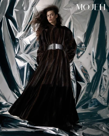 Fashion shot of a woman in a dark brown fur coat with a wide silver belt, posing in front of a shiny silver backdrop.