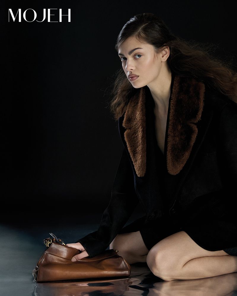 A brunette woman kneels and poses indoors holding a brown handbag. She wears a black jacket with a brown fur collar against a dark background.