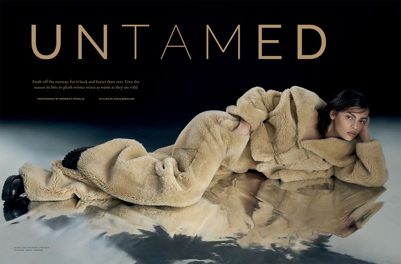 Fashion editorial shot of a woman in a fur coat lying on a reflective surface, with 'UNTAMED' text in gold letters above.