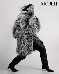 Monochrome fashion editorial image featuring a model wearing a textured faux-fur coat and tall black boots against a plain background.