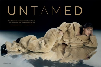 Model lying horizontally on a reflective surface wearing a plush beige faux fur jacket and matching trousers against a dark background.
