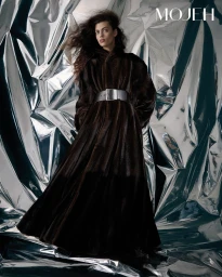Model with dark hair wearing a long dark belted coat against a reflective silver crinkled foil background
