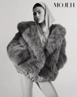 Monochrome fashion editorial featuring a model in a voluminous fur coat and sequined head covering against a plain background.
