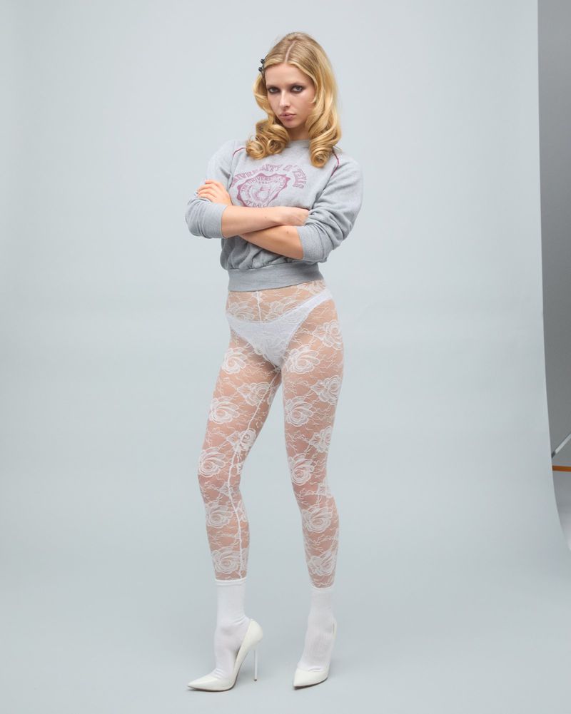 A blonde adult female model poses in a light grey sweatshirt, lace tights, and white heels against a light grey studio backdrop.