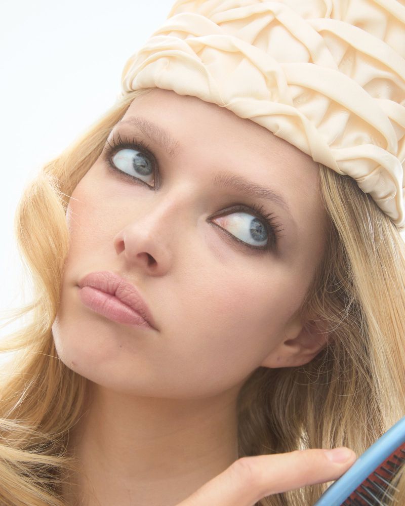 Close-up of a blonde adult female model wearing a textured cream-colored hat and holding a hairbrush against a plain white background.
