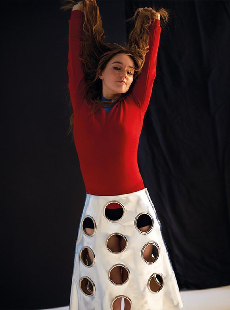 Young woman with long brown hair holds her hair up while wearing a red long-sleeve shirt and a white skirt with holes in a studio.