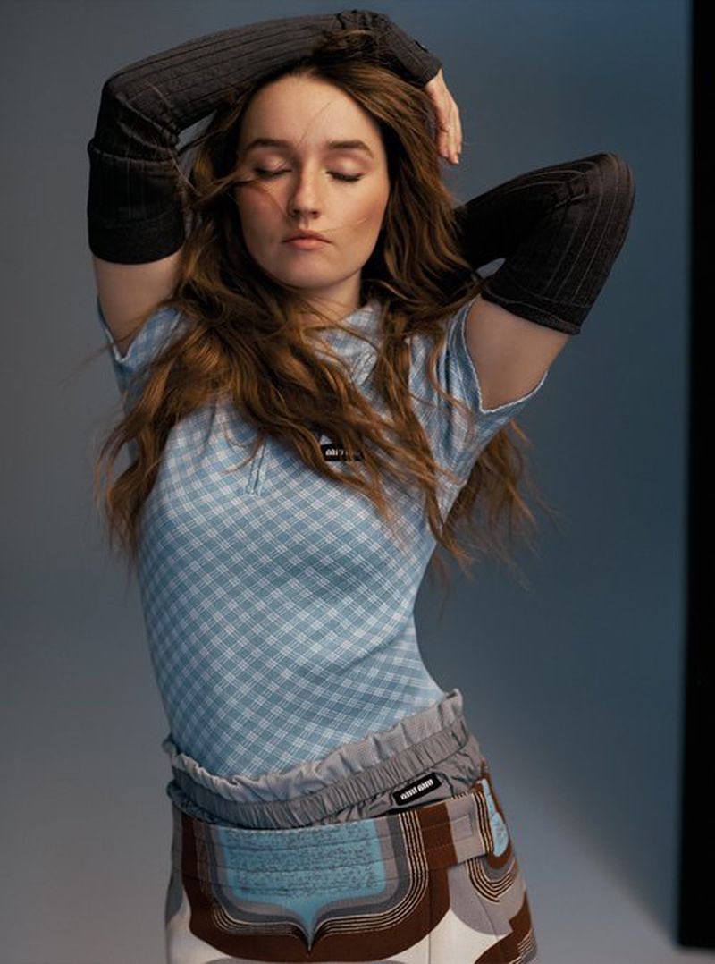 A young woman with long brown hair in a stylish checkered top and patterned skirt poses indoors with arms above her head.