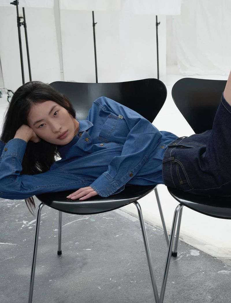 An Asian woman in a denim outfit reclines across two black chairs in a light-filled photo studio, posing casually for a fashion shot.