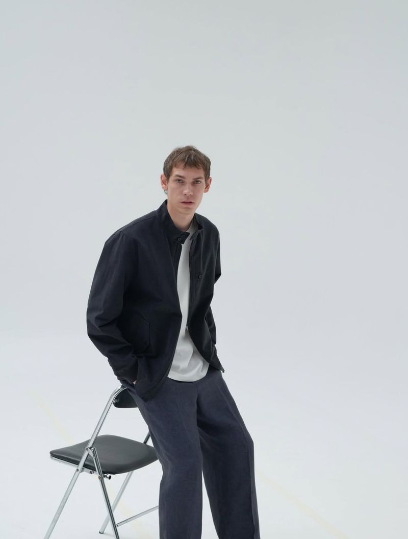 Adult male poses on a chair wearing a black jacket, t-shirt, and pants against a white backdrop in a studio setting. Fashion editorial.