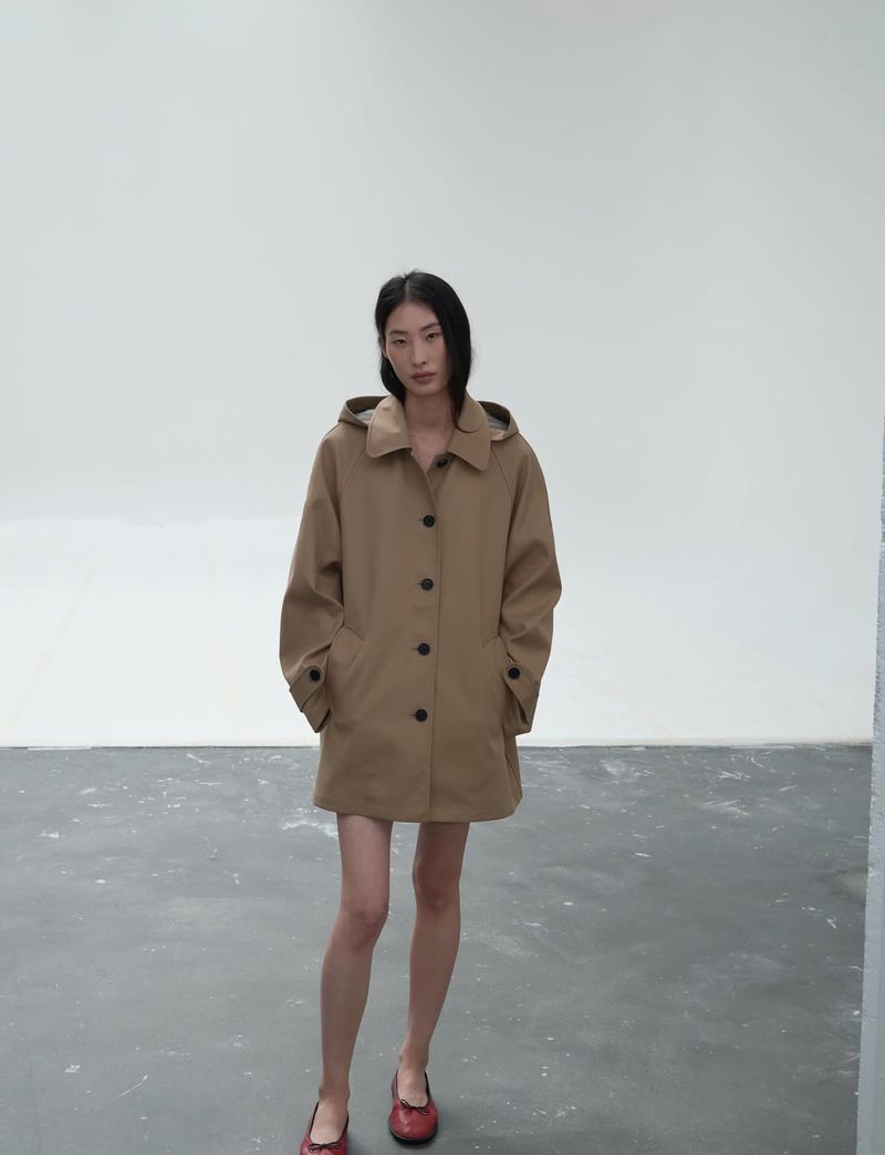 A slender Asian woman models a hooded, light brown trench coat and red Mary Jane shoes in a minimalist indoor studio setting with gray flooring.