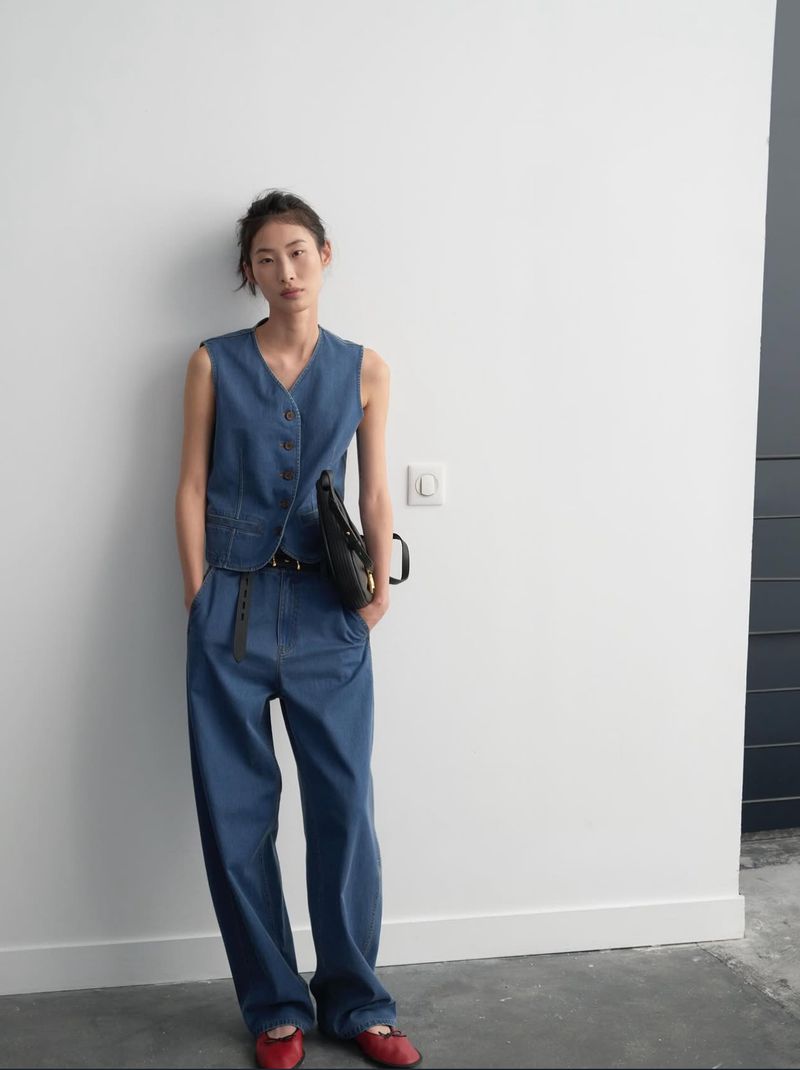 A woman poses in a blue denim vest and wide-leg pants, accessorized with a black belt and bag, and red ballet flats, against a white wall.