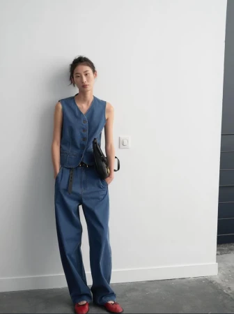 A woman poses in a blue denim vest and wide-leg pants, accessorized with a black belt and bag, and red ballet flats, against a white wall.