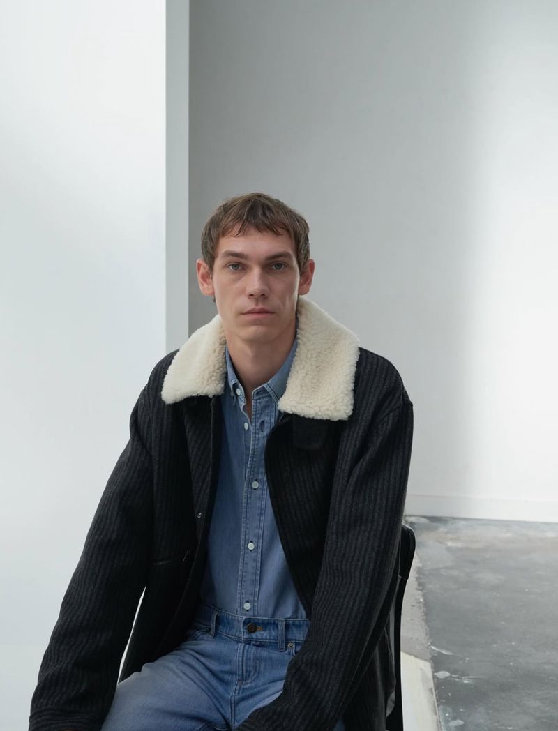 Adult man in a denim shirt and black corduroy jacket with white sherpa collar seated in a minimal studio with white walls and concrete floor.