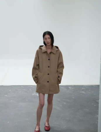 Model standing against a plain studio background wearing a tan hooded coat and red shoes
