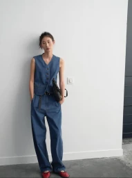 Model wearing a blue denim vest and matching trousers holding a black handbag against a white wall.