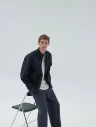 Man sitting on a metal folding chair against a white background wearing a dark jacket over a white top and dark trousers