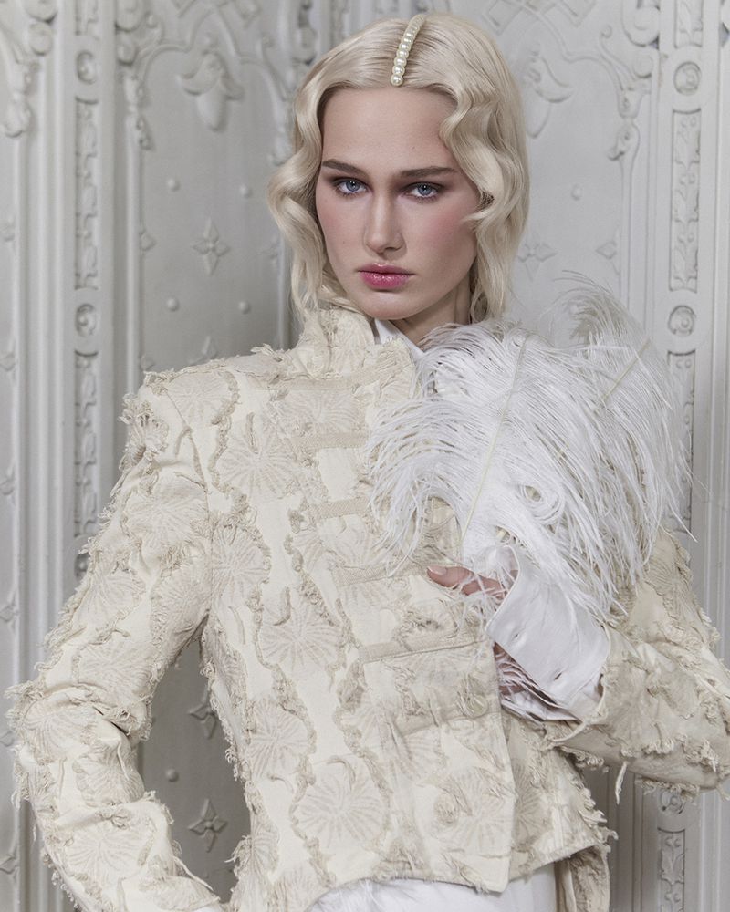 A blonde woman with an ornate white jacket poses in front of a detailed white wall, holding white feathers in her right hand.