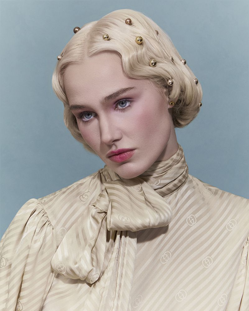 Portrait of a blonde woman with a beige, patterned, tied-neck blouse on a blue background, focusing on her face and stylish hair.