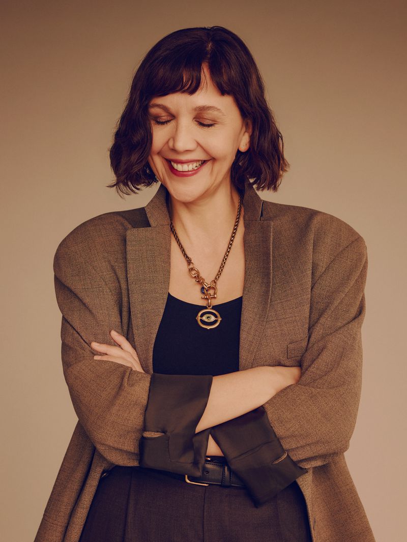 Smiling adult woman in a brown blazer with arms crossed, wearing a black top and a large pendant necklace against a neutral background.