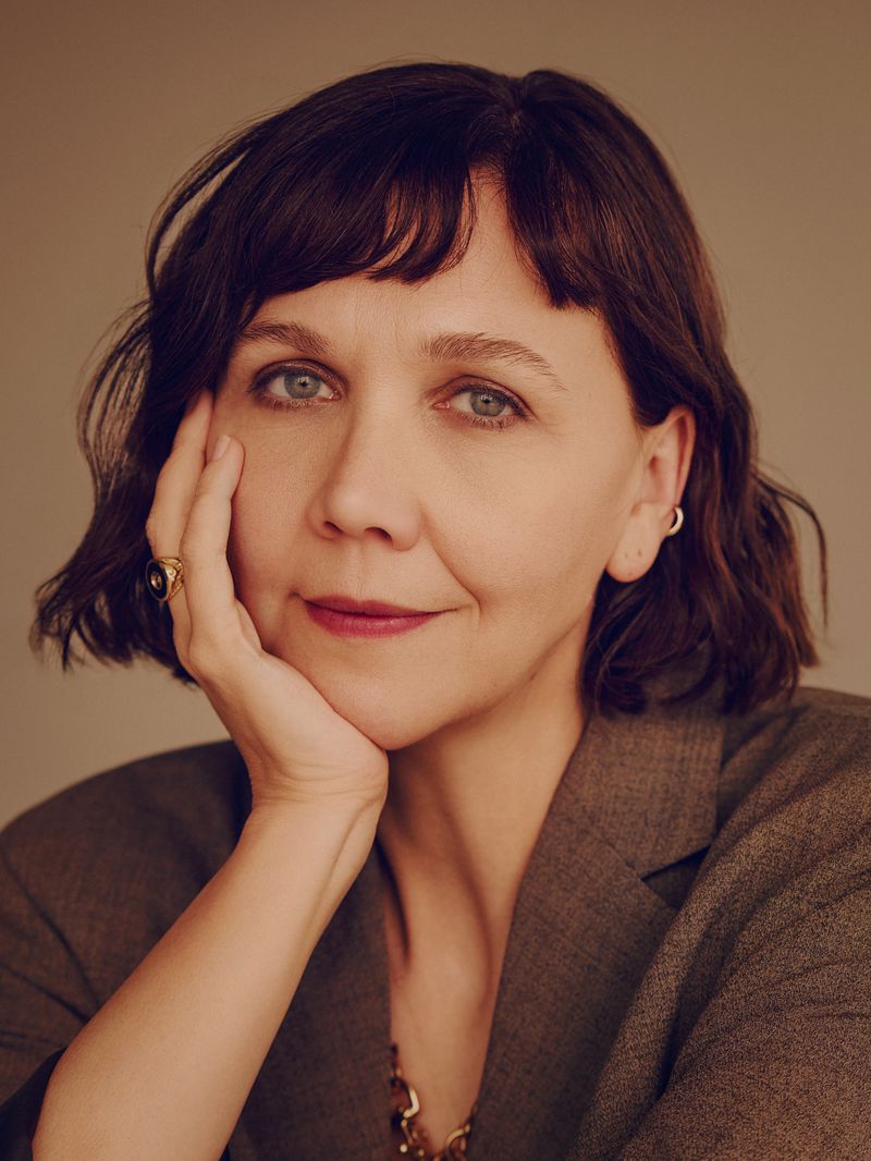 Close-up portrait of a woman with dark hair wearing a brown blazer and gold jewelry against a neutral background in a photo studio setting.