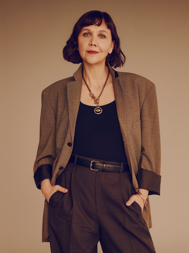 Adult woman with brown hair in a blazer and brown slacks posing in front of a tan background with hands in pockets.