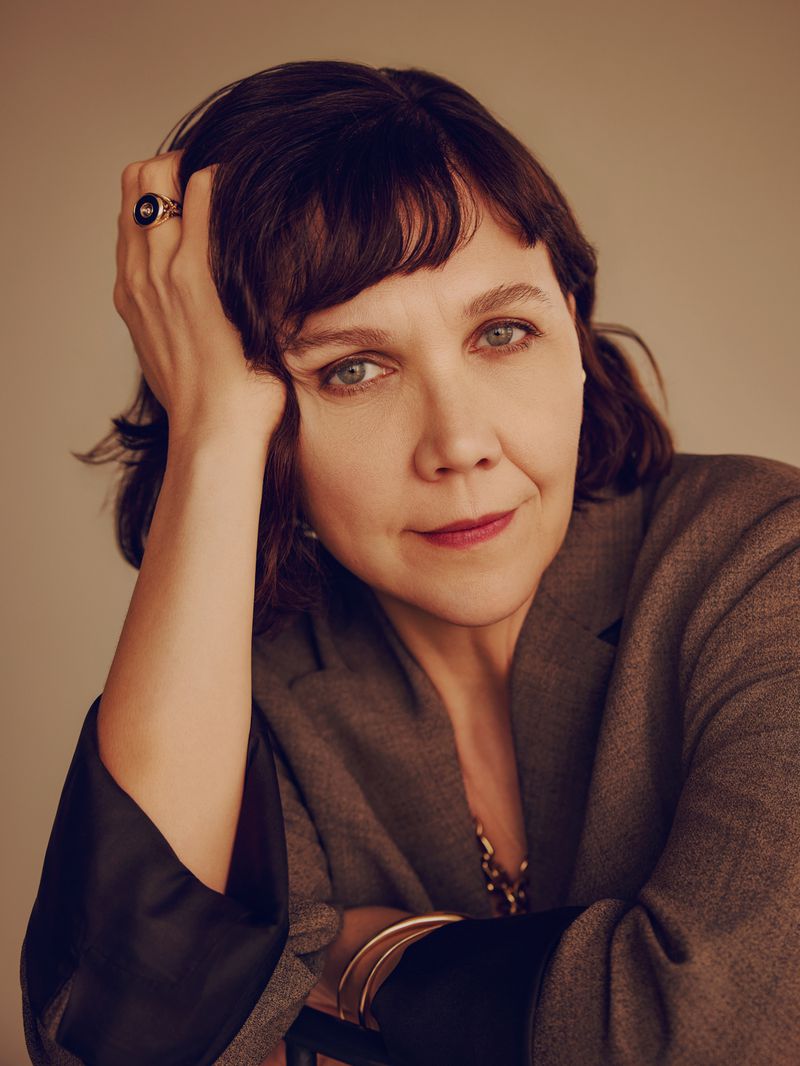 Portrait of a woman with dark hair and a grey blazer, resting her head on her hand, looking at the camera against a neutral background.