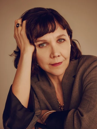 Portrait of Maggie Gyllenhaal resting hand on head wearing a grey blazer against a neutral backdrop