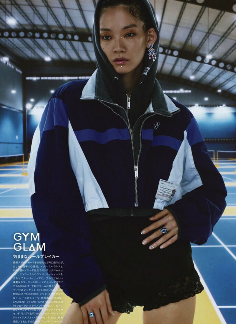 Fashion editorial shot of an Asian woman indoors wearing a cropped jacket, black shorts, and jewelry in a tennis court setting.