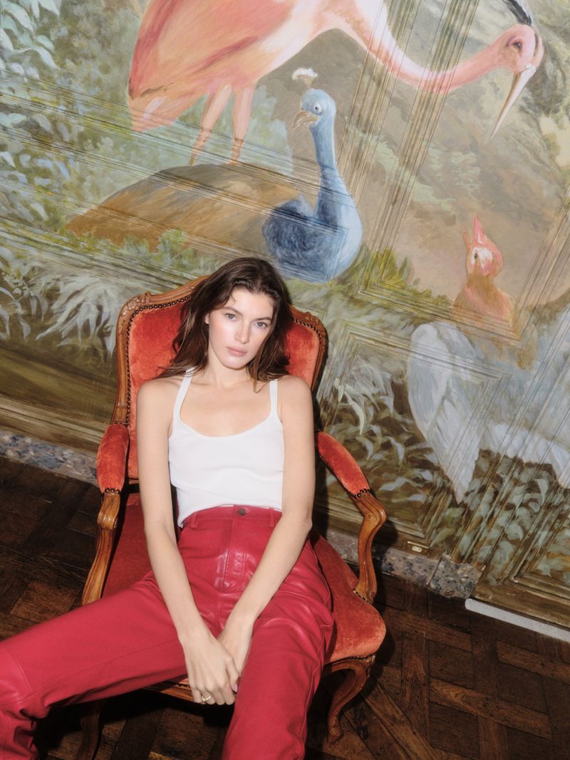 Woman with long dark hair poses in red pants and white top sitting in an ornate chair in front of a whimsical bird mural.