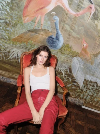 Woman with long dark hair poses in red pants and white top sitting in an ornate chair in front of a whimsical bird mural.