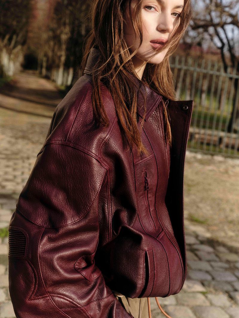 Portrait of a young adult female model wearing a textured, dark red leather jacket outdoors in a park-like setting on a sunny day.